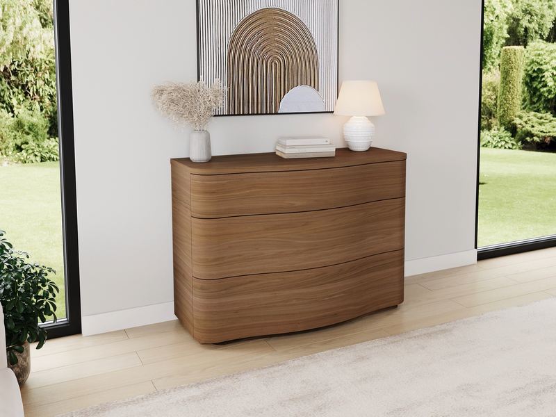 a wooden dresser with three drawers, a white vase with dried flowers, a white lamp, and two white books on top