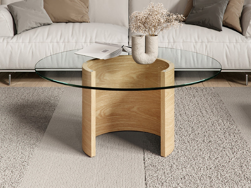 A close-up of a coffee table with a glass top and light-colored wooden base, with a vase of flowers and a book on top.